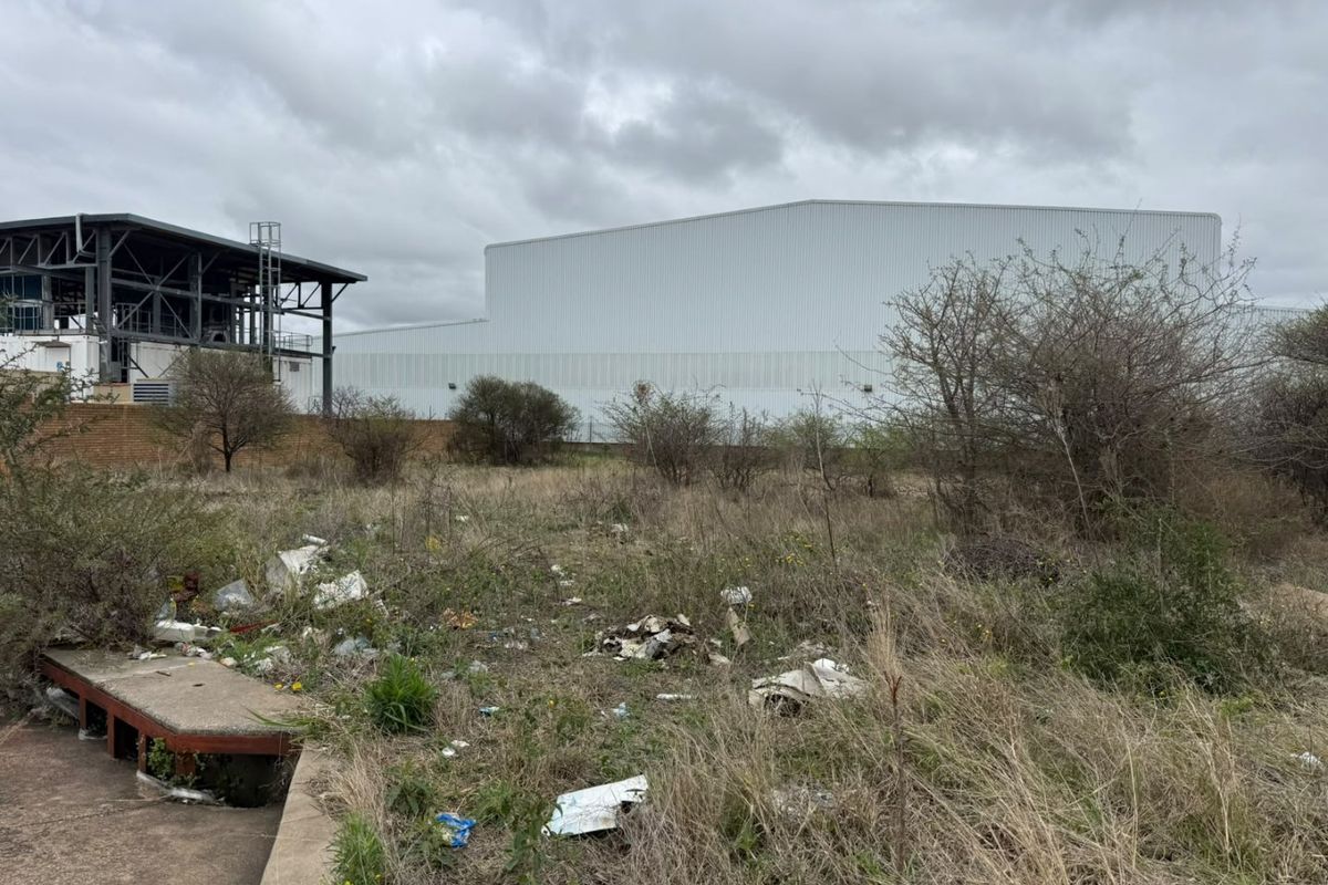 A view of a vacant residential plot featuring overgrown vegetation, debris, and a concrete slab, with an adjacent industrial structure in the background.