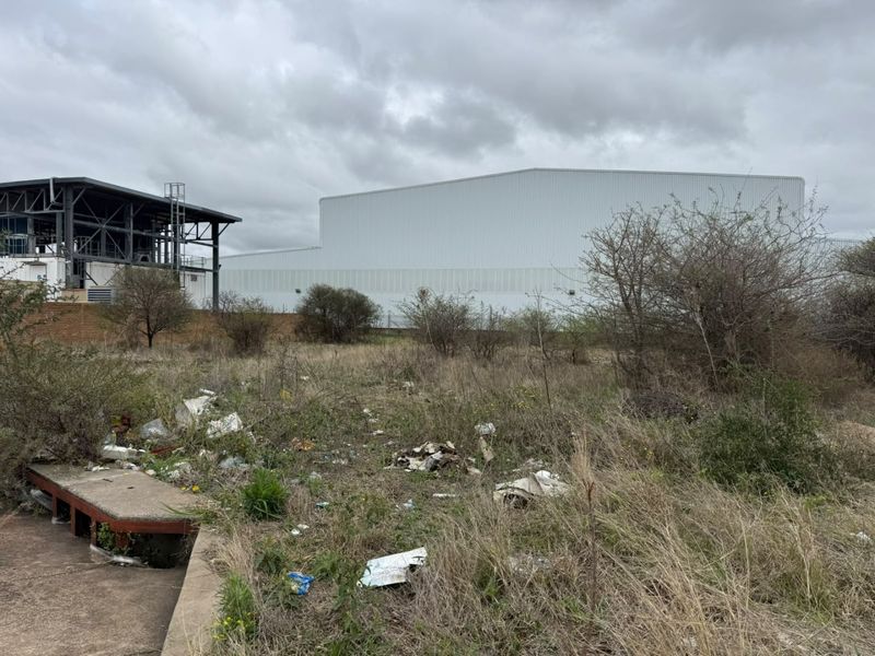 A view of a vacant residential plot featuring overgrown vegetation, debris, and a concrete slab, with an adjacent industrial structure in the background.
