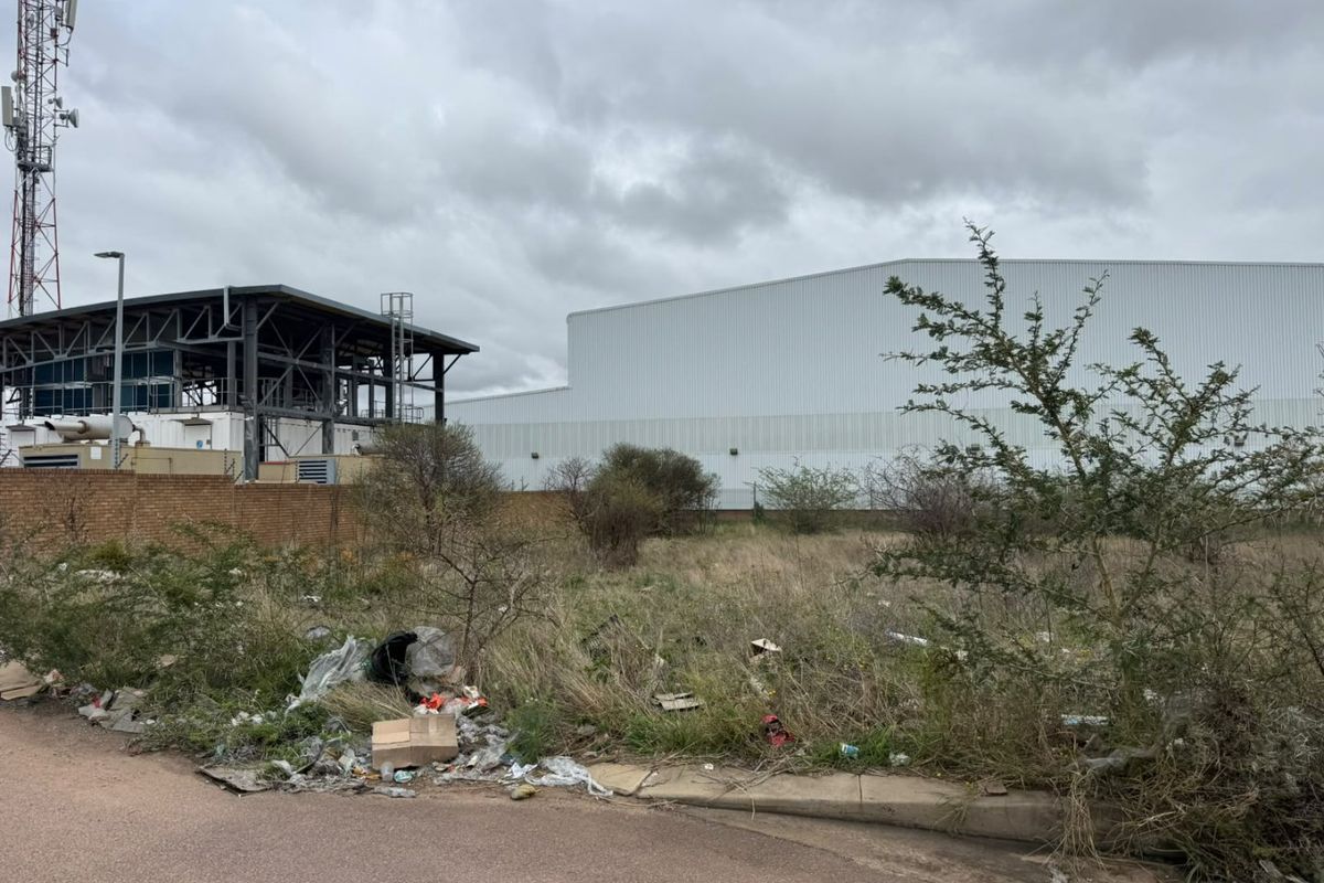 A vacant residential plot featuring a brick boundary wall, adjacent to industrial infrastructure and a paved road, currently requiring clearing of debris and vegetation.