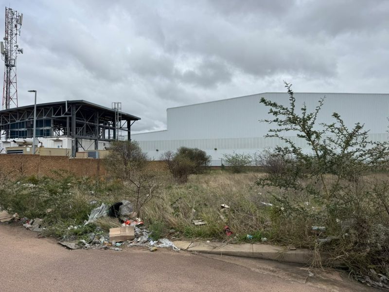 A vacant residential plot featuring a brick boundary wall, adjacent to industrial infrastructure and a paved road, currently requiring clearing of debris and vegetation.
