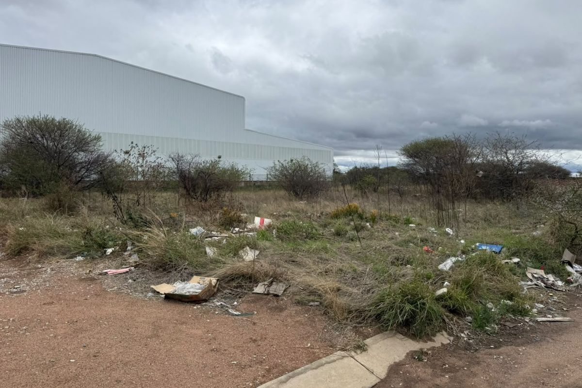 A vacant residential plot featuring native vegetation and accumulated debris, situated adjacent to a large industrial structure.