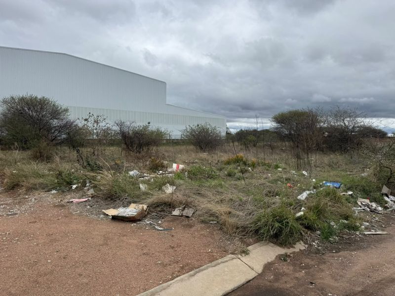 A vacant residential plot featuring native vegetation and accumulated debris, situated adjacent to a large industrial structure.