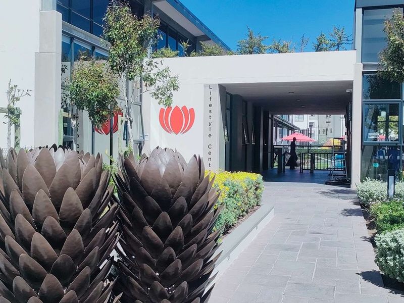 A well-maintained modern apartment complex entrance featuring a paved walkway, landscaped garden beds, and a covered access point to the lifestyle center.