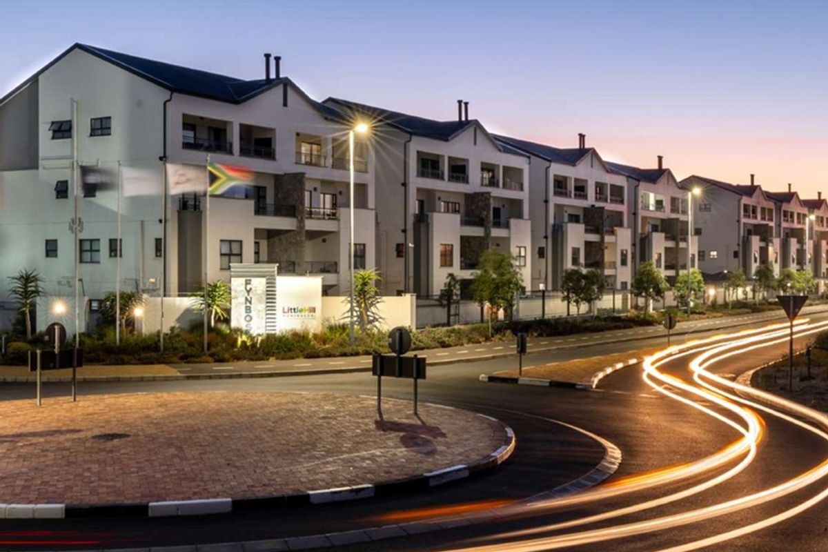 A twilight view of the Fynbos apartment complex, featuring modern multi-story architecture with balconies, situated adjacent to a paved roundabout and street infrastructure.