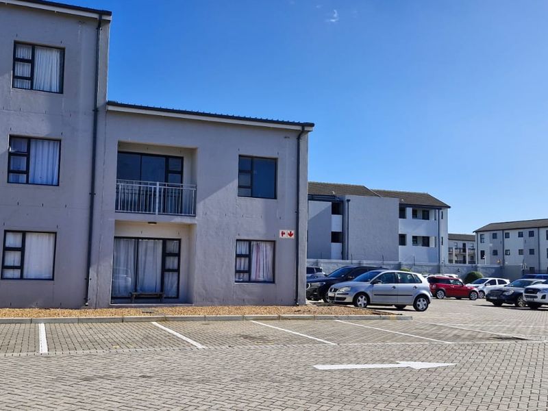 Exterior view of a multi-story apartment complex featuring paved parking, balconies with metal railings, and sliding glass doors.