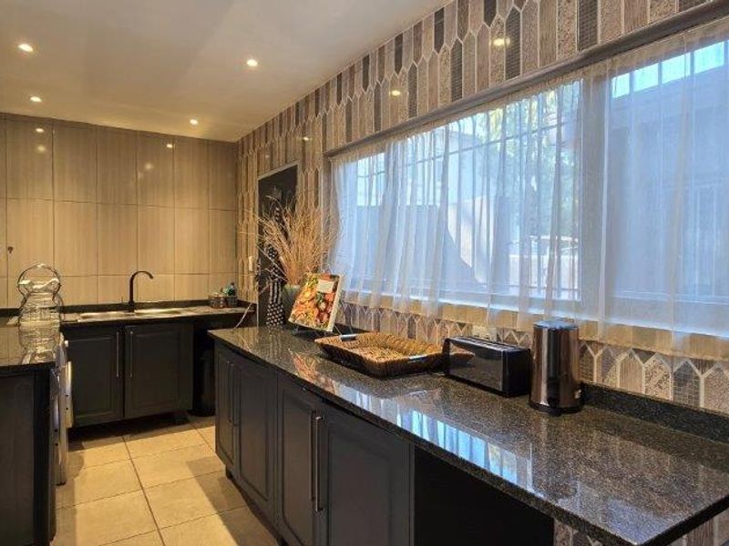 A well-appointed scullery featuring dark cabinetry, granite countertops, a stainless steel sink, and decorative tiled backsplash with ample natural light.