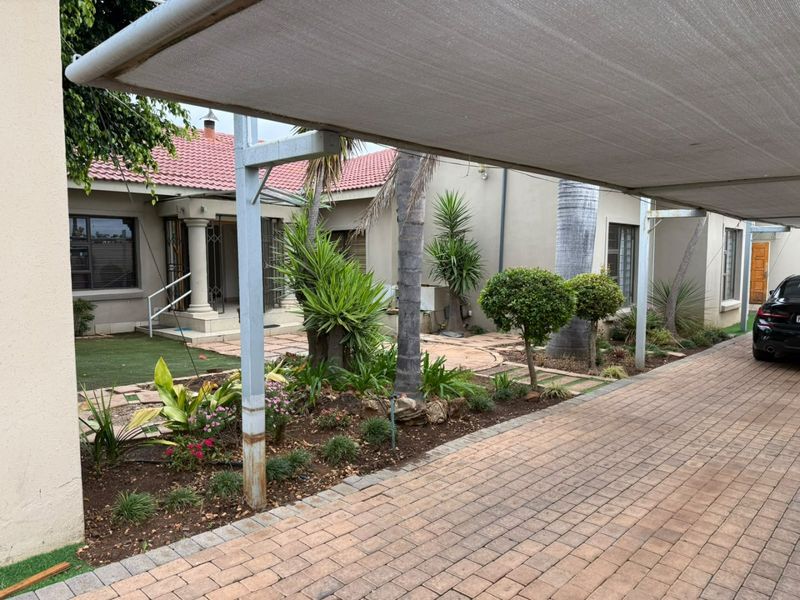 Front exterior view featuring a paved driveway, shade cloth carport, landscaped garden, and a tiled roof home with security gate entrance.