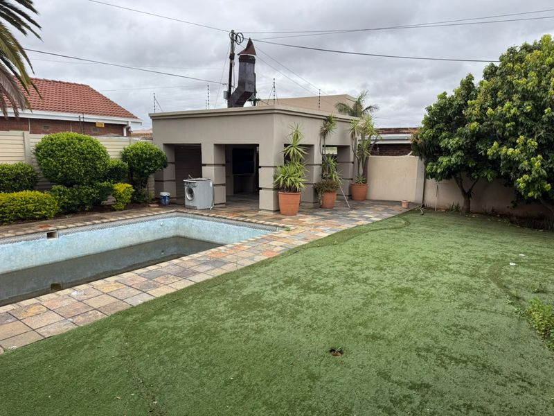 Outdoor pool area featuring a covered patio with built-in braai, stone paving, and an artificial lawn, providing a functional space for outdoor entertainment.