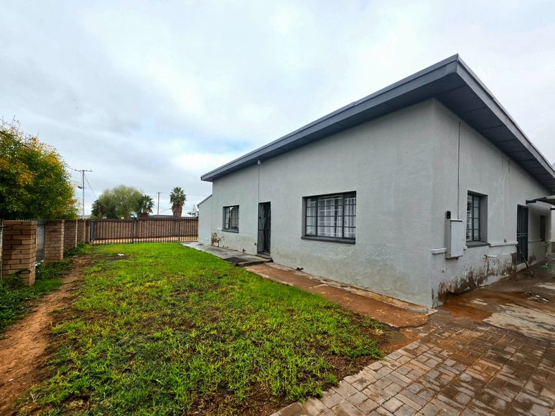 Rear exterior view of the property featuring a lawn, paved walkway, and boundary wall, showing signs of exterior wall maintenance requirements.