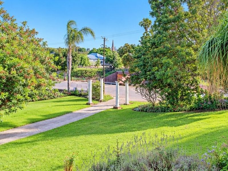 A well-maintained garden featuring a paved walkway leading through decorative pillars, surrounded by lush green lawns and mature trees.