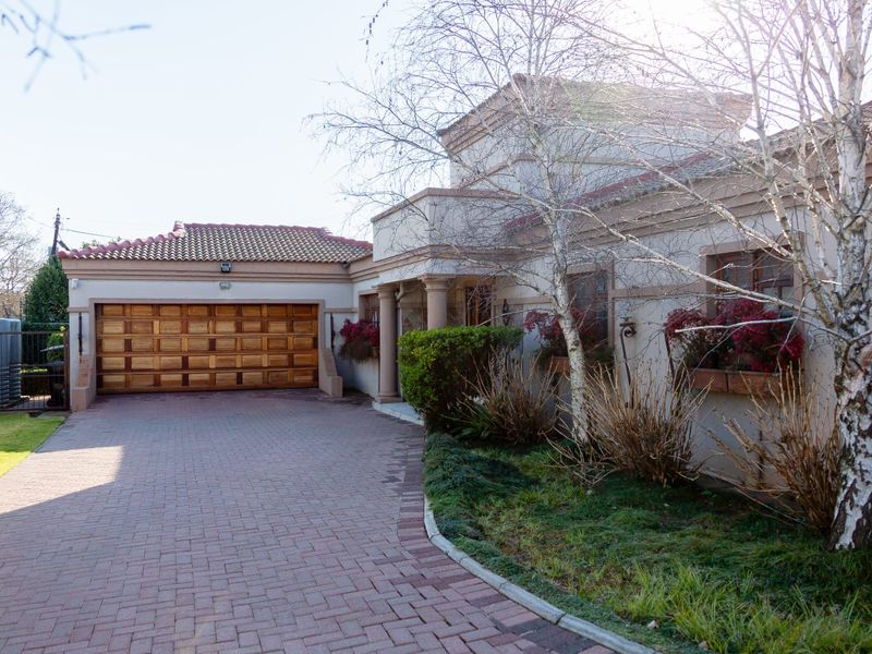 Front exterior view of the house featuring a paved driveway, a wooden garage door, and a landscaped garden area with mature trees.