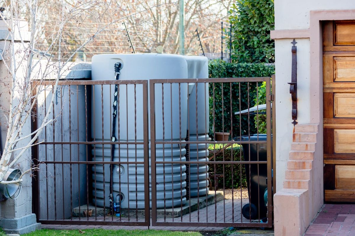 A side yard area featuring a large water storage tank secured behind a metal gate, adjacent to a wooden garage door.