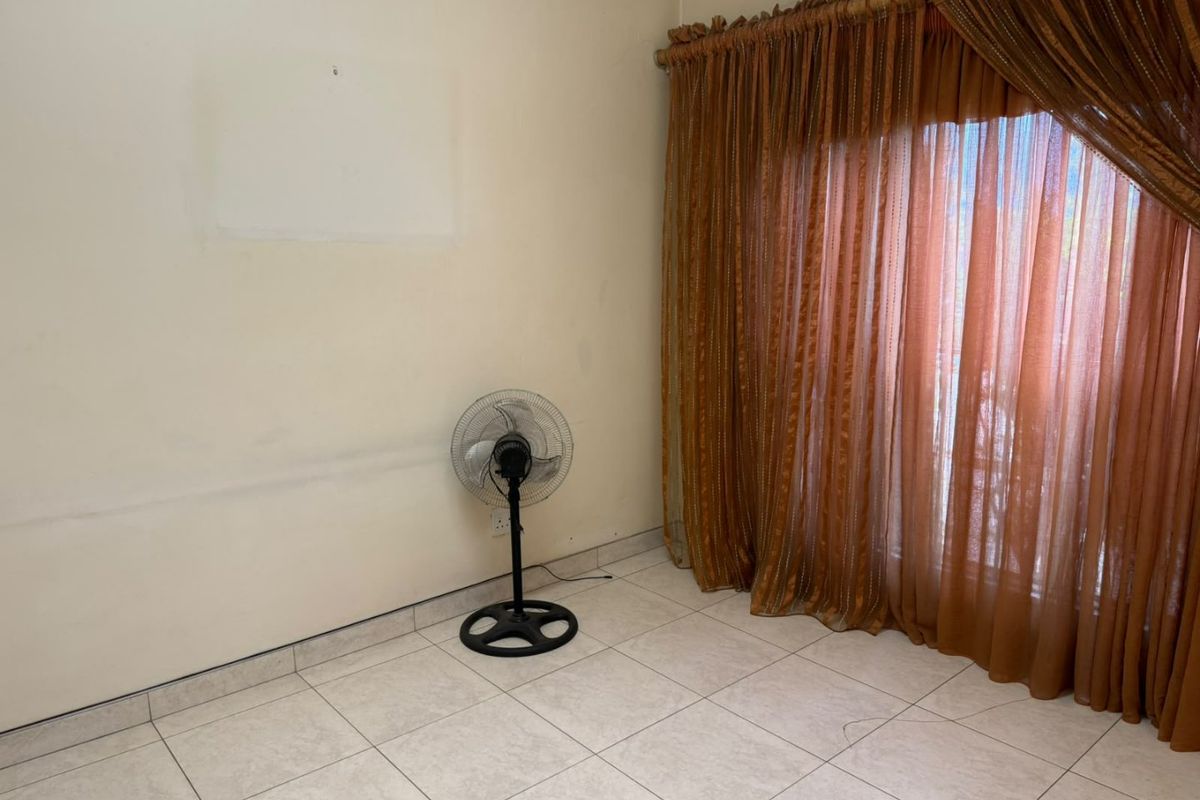 An empty living area featuring tiled flooring and large window treatments, currently showing signs of wear on the walls.