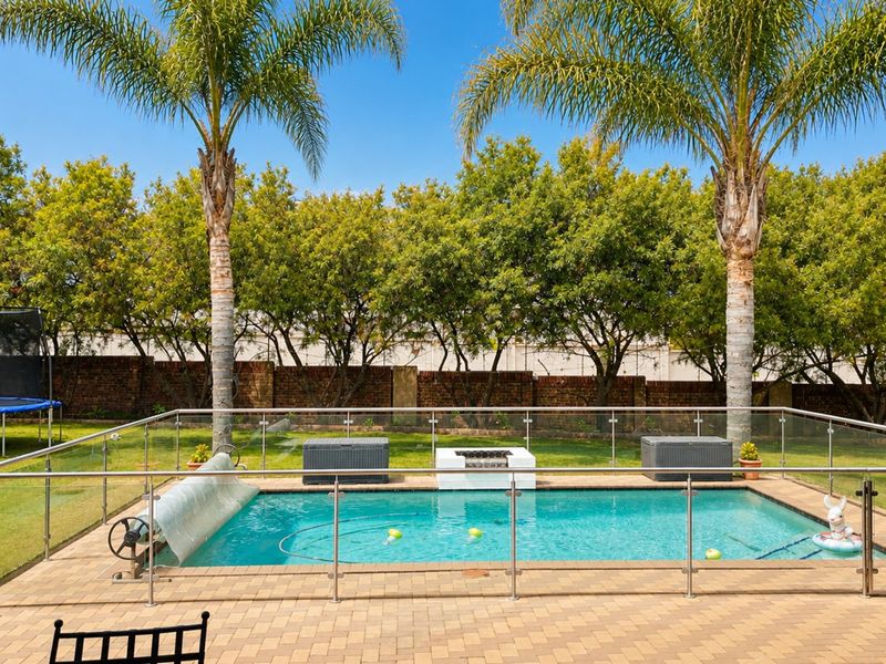 A well-maintained outdoor pool area featuring a paved surround, glass safety fencing, and a built-in fire pit set against a landscaped garden backdrop.