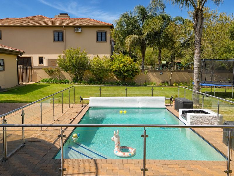 A well-maintained backyard featuring a swimming pool with glass fencing, a paved patio area, and a spacious lawn.