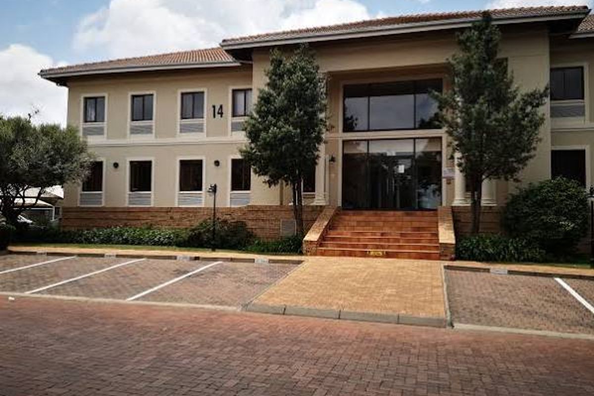 A professional commercial office building exterior featuring a paved parking area, brick facade, and a prominent glass entrance with steps.
