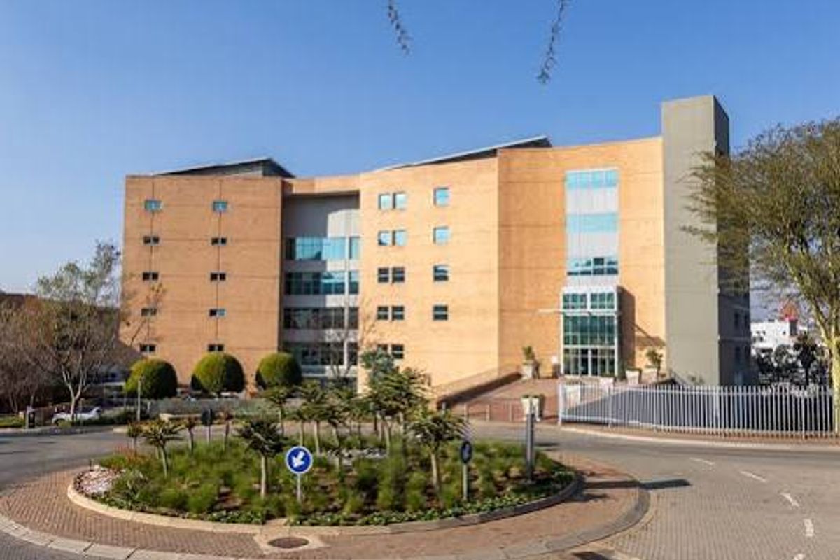 A professional view of a multi-story commercial office building featuring brick and glass architecture, situated behind a landscaped roundabout and paved entrance area.