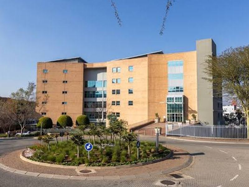 A professional view of a multi-story commercial office building featuring brick and glass architecture, situated behind a landscaped roundabout and paved entrance area.