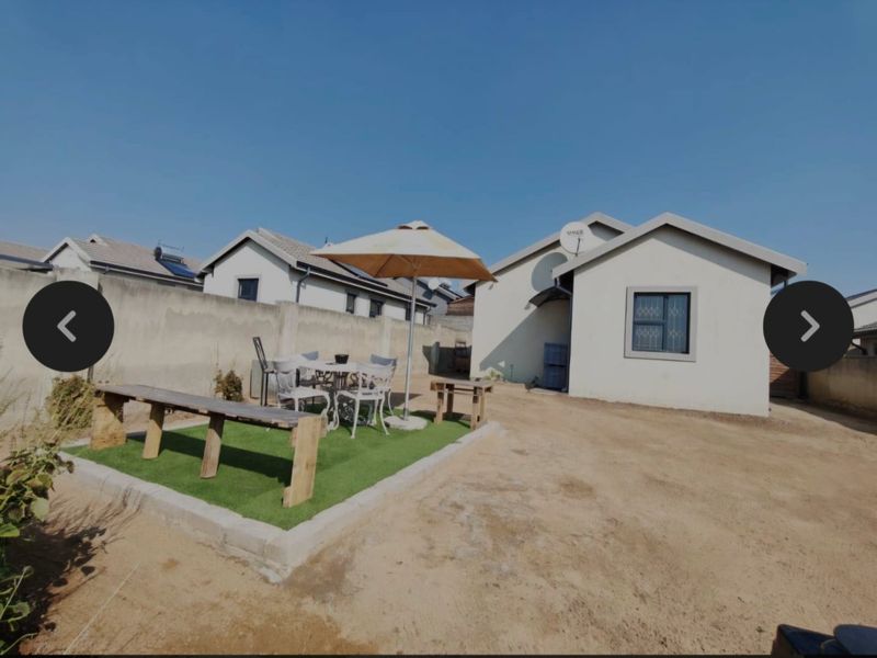 Rear exterior view of the property featuring a raised artificial turf area, perimeter wall, and satellite dish, set against an unpaved sandy yard.