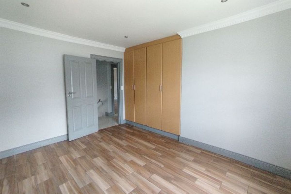 A bright, empty bedroom featuring wood-look tiled flooring, built-in cupboards, and recessed downlighting with decorative coving.