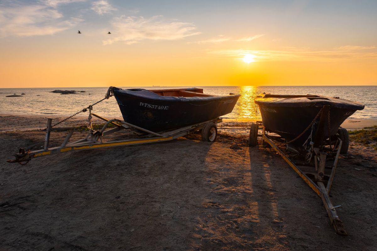 A scenic sunset view over the ocean from the shoreline, featuring boats on trailers parked on the sand.