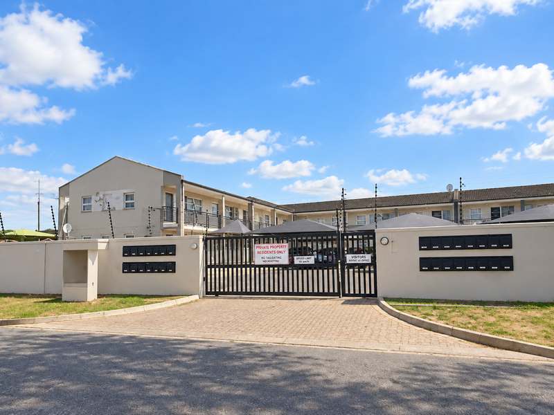 Front entrance of the apartment complex featuring a secure gated access point, paved driveway, perimeter wall with electric fencing, and communal post boxes.