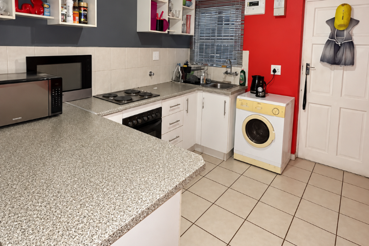 Functional kitchen featuring built-in cabinetry, laminate countertops, an electric hob, oven, and integrated laundry plumbing, finished with tiled flooring and a bold accent wall.