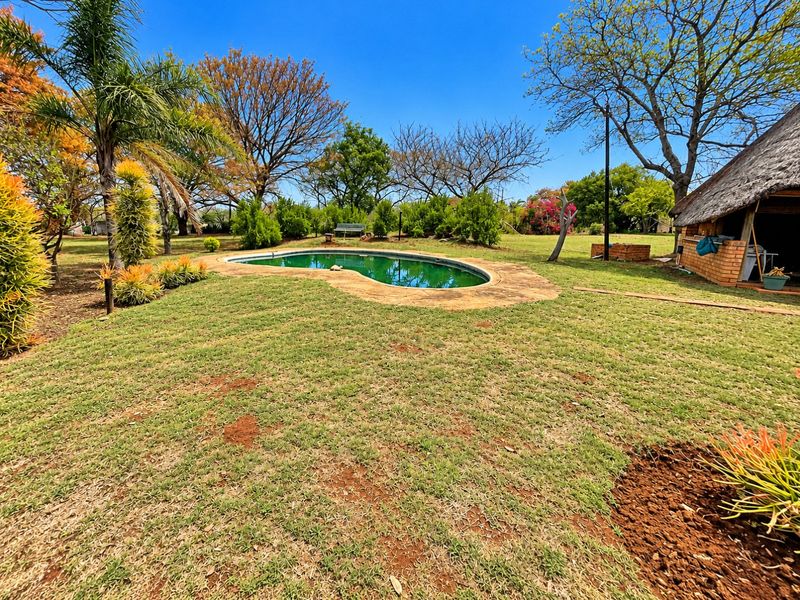 A spacious outdoor area featuring a swimming pool set within a large garden, accompanied by a rustic thatched-roof structure.