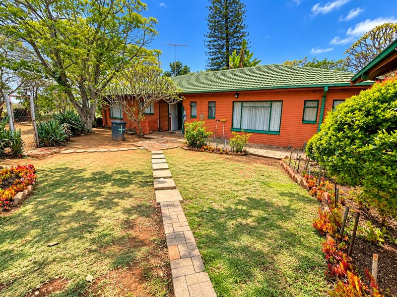 A rear view of a single-story brick house featuring a green tiled roof, paved garden walkway, and established landscaping.