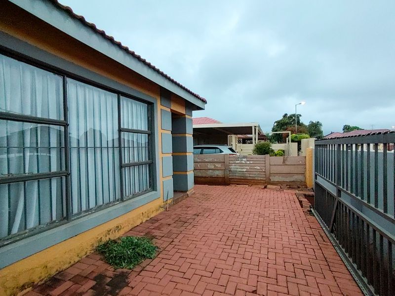 Front exterior view showing a paved driveway, metal sliding gate, and precast boundary wall with visible wear and maintenance requirements.