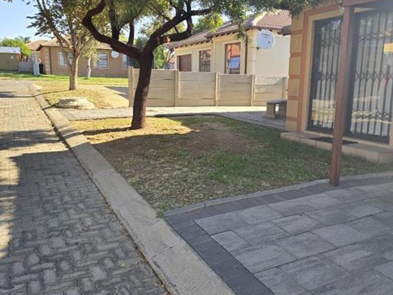 Front exterior view of the property featuring a paved driveway, security gate, and established garden area within a residential complex.