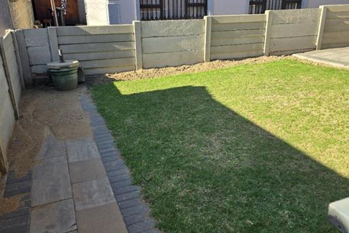 A rear garden area featuring a lawn, paved walkway, and precast concrete boundary wall, adjacent to the main house structure.