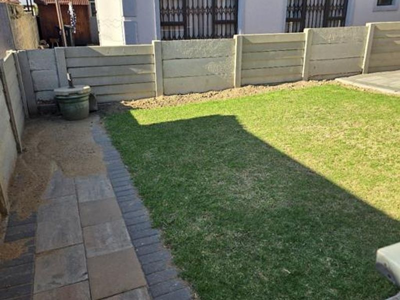 A rear garden area featuring a lawn, paved walkway, and precast concrete boundary wall, adjacent to the main house structure.