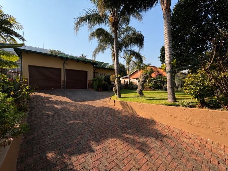 Front view of the property featuring a paved driveway, double garage, and a well-maintained garden with mature palm trees.