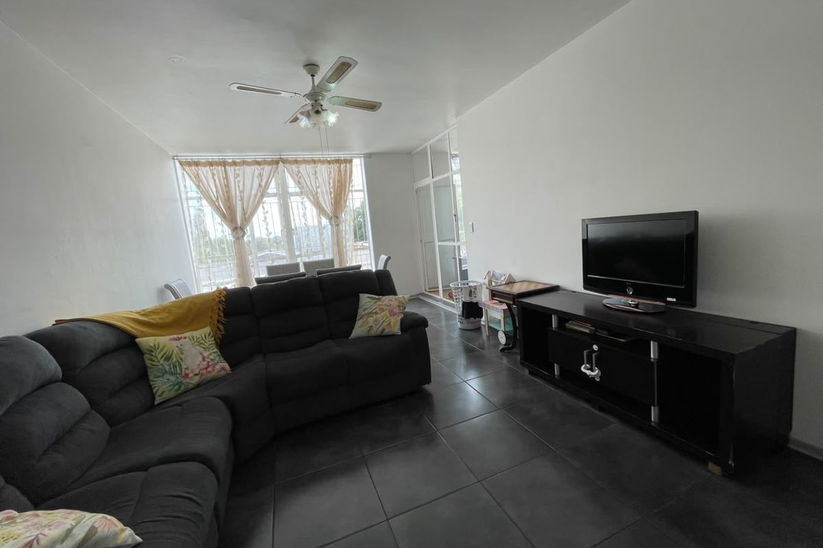 A living room featuring dark tiled flooring, a ceiling fan, and large windows providing natural light, with access to an adjacent balcony area.