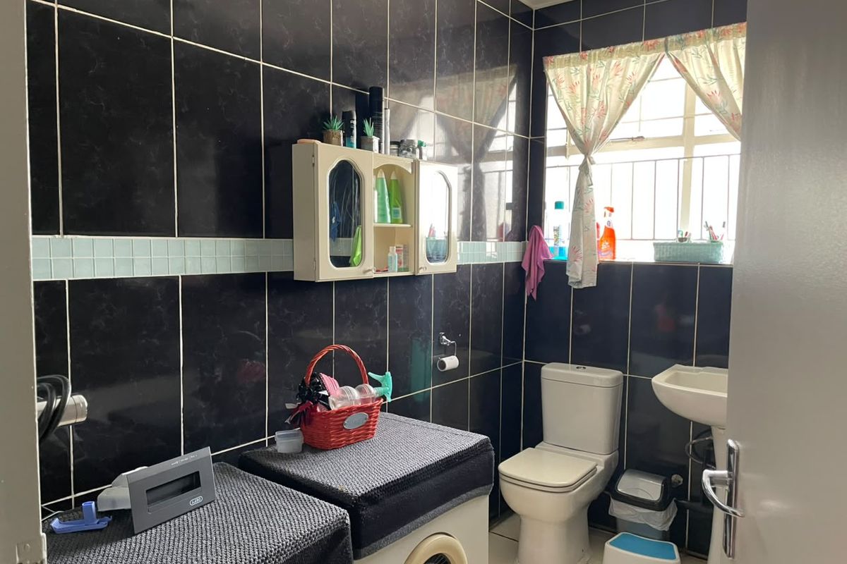 A functional bathroom featuring black wall tiling, a pedestal basin, a standard toilet, and a wall-mounted storage cabinet with security bars on the window.