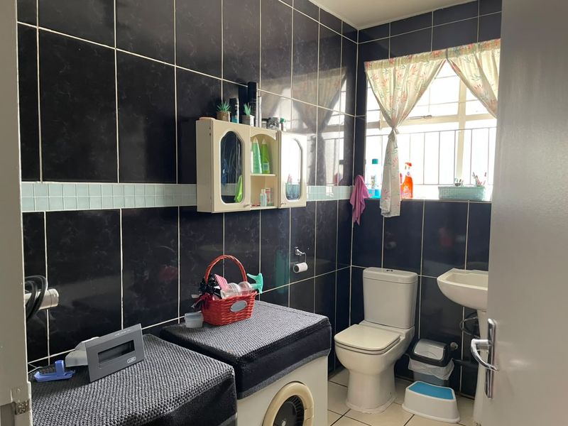 A functional bathroom featuring black wall tiling, a pedestal basin, a standard toilet, and a wall-mounted storage cabinet with security bars on the window.
