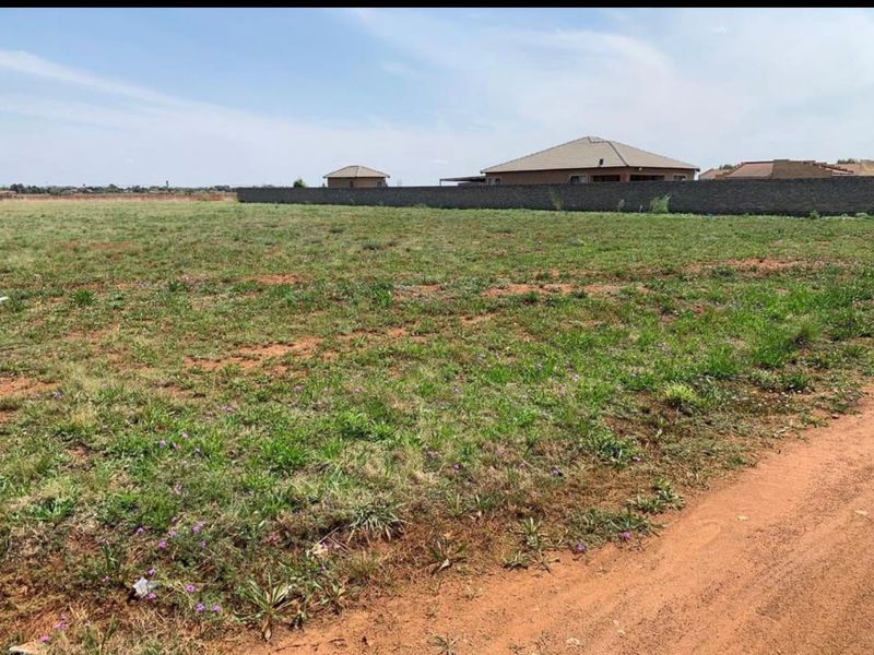 A vacant residential plot of land featuring natural grass cover, a perimeter boundary wall, and access via an unpaved road.
