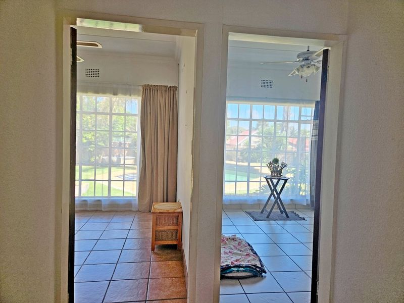 A view from a hallway looking into two adjacent rooms, both featuring tiled floors, ceiling fans, and large windows overlooking a garden and pool area.