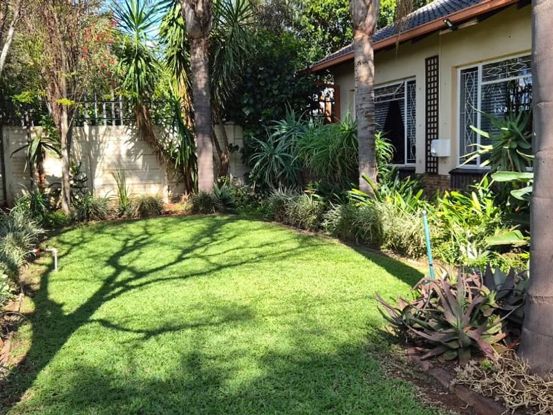 A well-maintained residential garden featuring a lush green lawn, mature trees, and established landscaping alongside the side elevation of the house.
