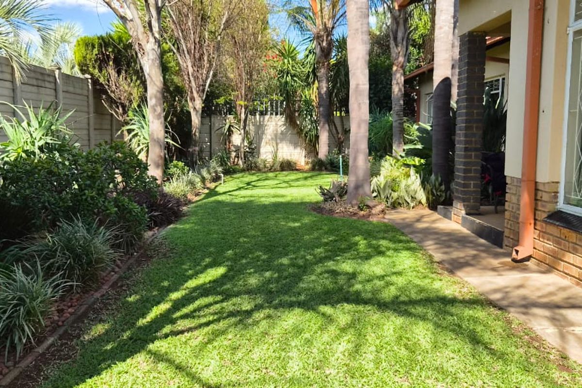 A well-maintained rear garden featuring a lush lawn, mature trees, and a paved walkway adjacent to the brick-clad exterior of the house.