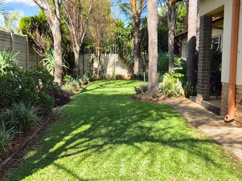 A well-maintained rear garden featuring a lush lawn, mature trees, and a paved walkway adjacent to the brick-clad exterior of the house.