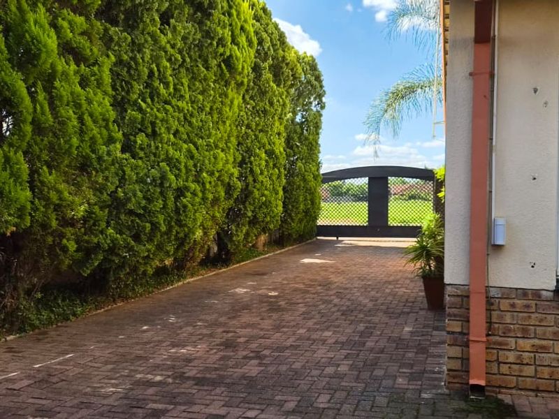 A paved driveway leading to a secure metal gate, flanked by mature hedging and a brick-accented exterior wall.