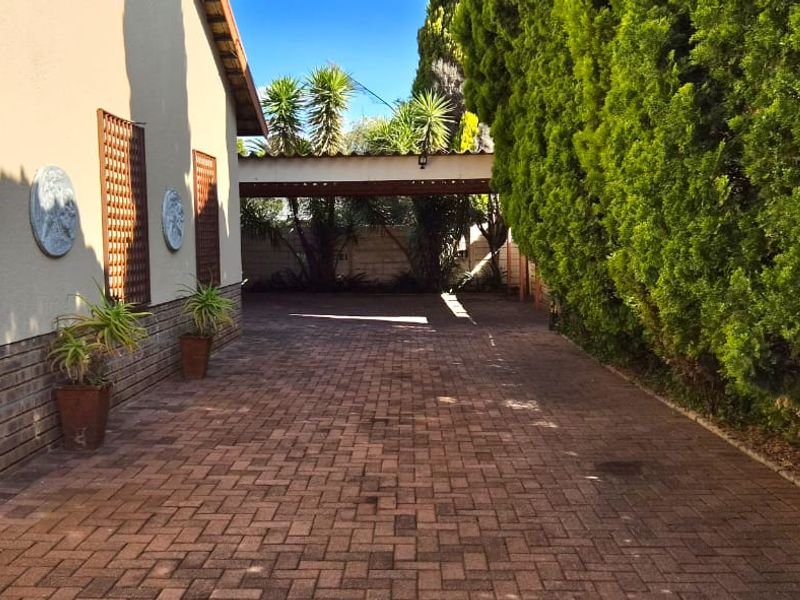A well-maintained rear exterior area featuring a brick-paved driveway, a covered carport, and established perimeter hedging.