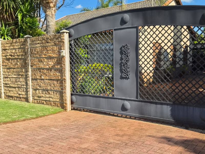 A secure front entrance featuring a decorative metal driveway gate, solid brick boundary walling, and a neatly paved driveway.