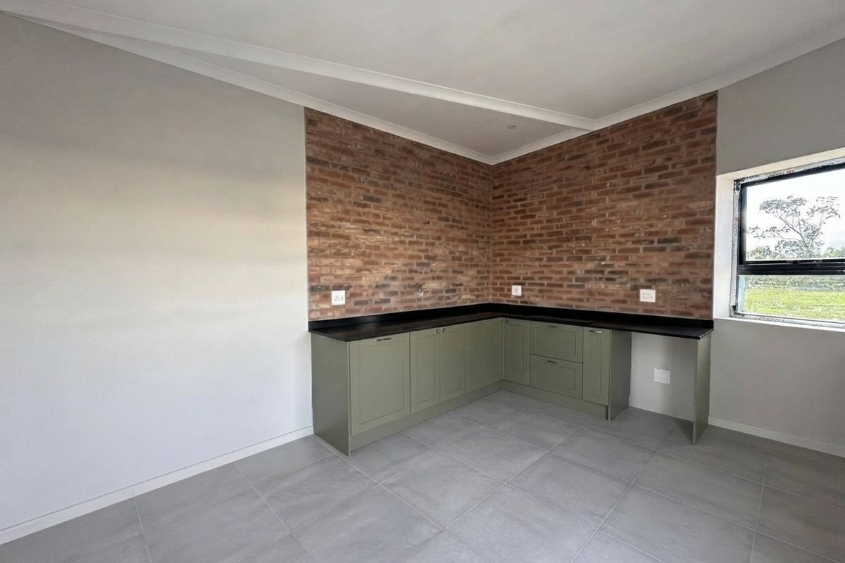 A modern, empty kitchen area featuring olive green cabinetry, dark stone countertops, and a striking exposed brick feature wall with large format floor tiles.
