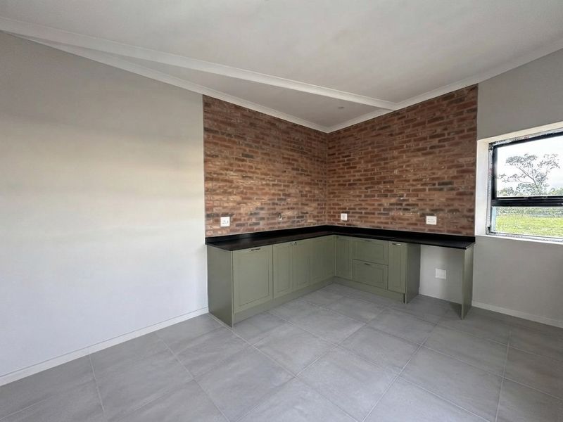 A modern, empty kitchen area featuring olive green cabinetry, dark stone countertops, and a striking exposed brick feature wall with large format floor tiles.