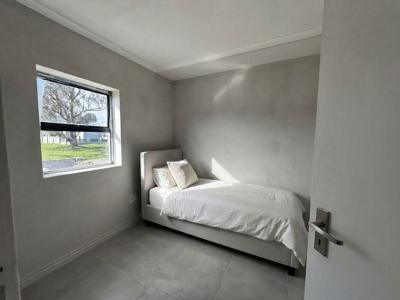 A compact bedroom featuring neutral grey wall finishes, large format floor tiles, and an aluminium-framed window providing natural light.