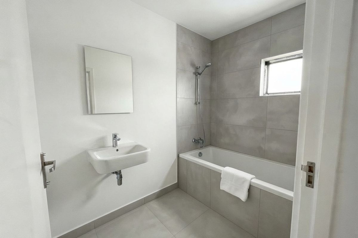 A modern, minimalist bathroom featuring a wall-mounted basin, a bathtub with shower attachment, and neutral grey tiling throughout.
