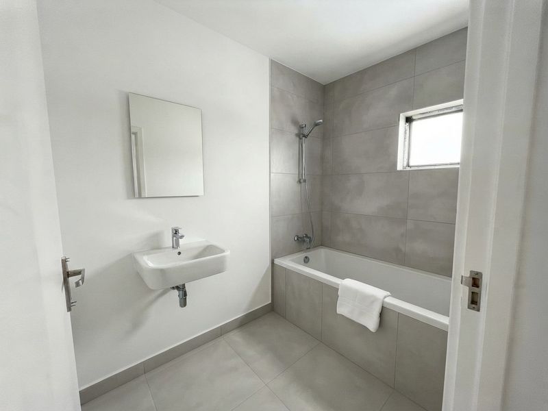 A modern, minimalist bathroom featuring a wall-mounted basin, a bathtub with shower attachment, and neutral grey tiling throughout.